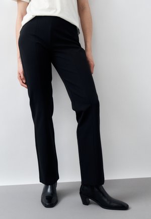 Osoba stojaca pri bielej stene a sivom podlahe, oblečená v priamych čiernych nohaviciach a čiernych členkových čižmách na podpätku.