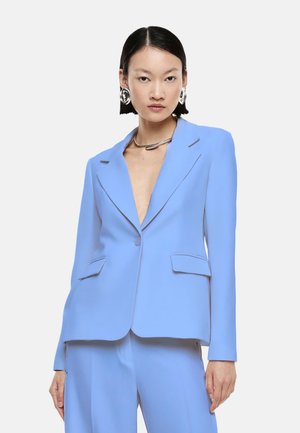 Donna che indossa un completo sartoriale azzurro chiaro con blazer scollo a V profondo, pantaloni a gamba larga, collana d'argento e grandi orecchini a cerchio d'argento su sfondo bianco.