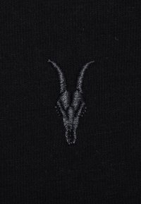 Tissu noir avec un petit crâne de chèvre argenté brodé en détail, présentant de longues cornes courbées, centré sur la surface.