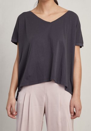 Vrouw draagt een losse, donkergrijze, korte mouw V-hals top en een lichtbeige, geplisseerde, broek met hoge taille, staand tegen een effen achtergrond.