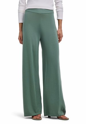 Personne portant un pantalon large vert sauge, un haut blanc à manches longues et des tongs, debout devant un fond blanc.
