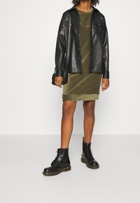 Chaqueta de camisa de cuero negra sobre un vestido de terciopelo verde oliva. Botas negras con cordones y acentos amarillos. Modelo sosteniendo un pequeño bolso.