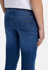 Jeans in denim blu scuro con vestibilità slim, dotati di tasche posteriori e dettagli di cucitura sottili. Il tessuto ha una texture liscia e una leggera scoloritura.