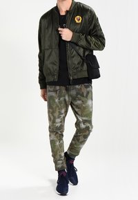 Olivenfarbene Bomberjacke mit einem Aufnäher, schwarzes T-Shirt, Camouflage-Hose und dunkle Sneakers. Zu den Merkmalen gehört eine Reißverschlusstasche an der Hose.
