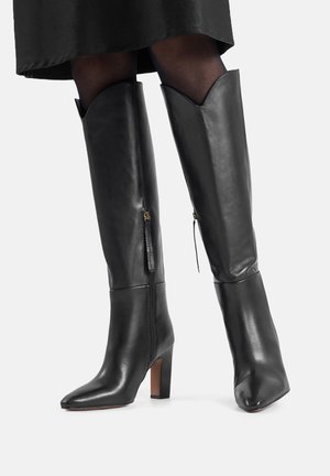 Schwarze kniehohe Lederstiefel mit spitzer Kappe und Blockabsatz, ausgestattet mit einem seitlichen Reißverschluss und einem gezackten oberen Rand als Detail.