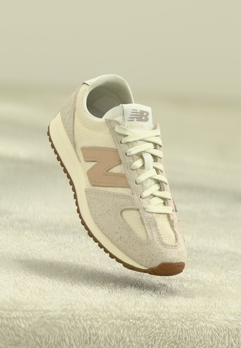 Baskets New Balance crème et beige avec un mélange de textures en maille et en daim, logo marron clair et lacets blancs, présentées sur un fond texturé doux.