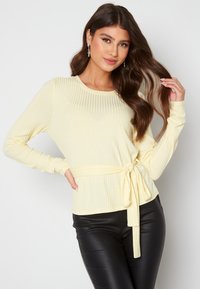 Top a maniche lunghe ribbed giallo chiaro con scollatura rotonda e cintura annodata in vita. Tessuto morbido, abbinato a pantaloni neri aderenti.