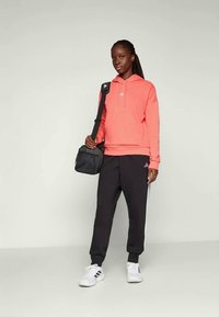 Mulher vestindo um hoodie corais, calças de treino pretas da Adidas, tênis brancos e carregando uma bolsa de ombro preta, de pé contra um fundo liso.