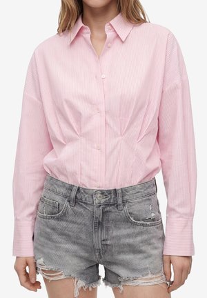 Mujer que lleva una camisa de botones de rayas rosas con mangas largas y pantalones cortos vaqueros desgastados grises de talle alto.