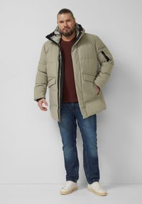 Beżowa kurtka puchowa z zamkiem z przodu, dużymi kieszeniami i czarną podszewką kaptura, zestawiona z ciemnymi dżinsami i białymi trampkami.