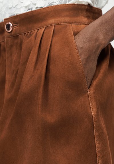 Χέρι στην τσέπη καφέ δερμάτινης φούστας με υψηλή μέση, πιέτες, ζώνη με κουμπιά και εμφανείς ραφές.