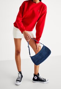 Personne portant un sweat-shirt rouge, une mini-jupe blanche et des baskets montantes noires, tenant un sac épaule en denim bleu sur un fond blanc.