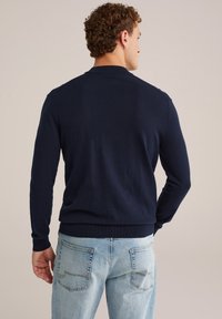 Man som vänder ryggen mot kameran iförd en marinblå långärmad tröja och ljusblå denimjeans mot en enkel bakgrund.