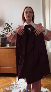 Ohlapna bordo jakna z zarezanimi ovratniki, zapiranjem na en gumb in mehko teksturo, držana pred svetlim ozadjem z lesenim pohištvom.