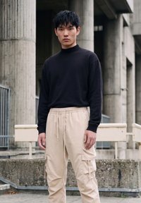 Junger Mann mit schwarzen Haaren, der ein schwarzes Langarmshirt und beige Cargo-Hosen trägt, steht im Freien nahe Betonpfeilern.