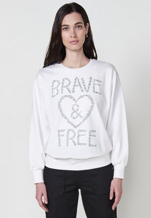 Biele mikina s dlhými rukávmi s veľkým potlačeným dizajnom, ktorý hovorí "BRAVE ❤️ FREE" v strieborných štofoch. Hladká textúra, voľný strih.