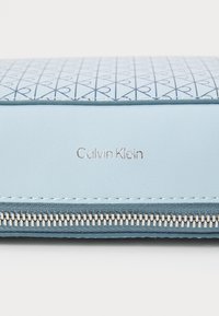 Ljust blå plånbok med ett mönstrat lock som har sammanlänkade hjärtan, silverfärgad dragkedja och präglad "Calvin Klein"-logotyp på en slät sektion.