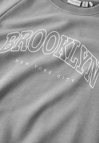 Sudadera gris con "BROOKLYN" en letras grandes blancas y "NEW YORK CITY" en texto más pequeño, con un cuello acanalado y una textura suave.