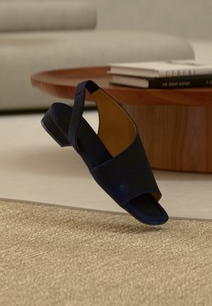 Enkele marineblauwe open-teen slingback sandaal zwevend boven een gestructureerd beige vloerkleed, met een houten salontafel en gestapelde boeken op de achtergrond.