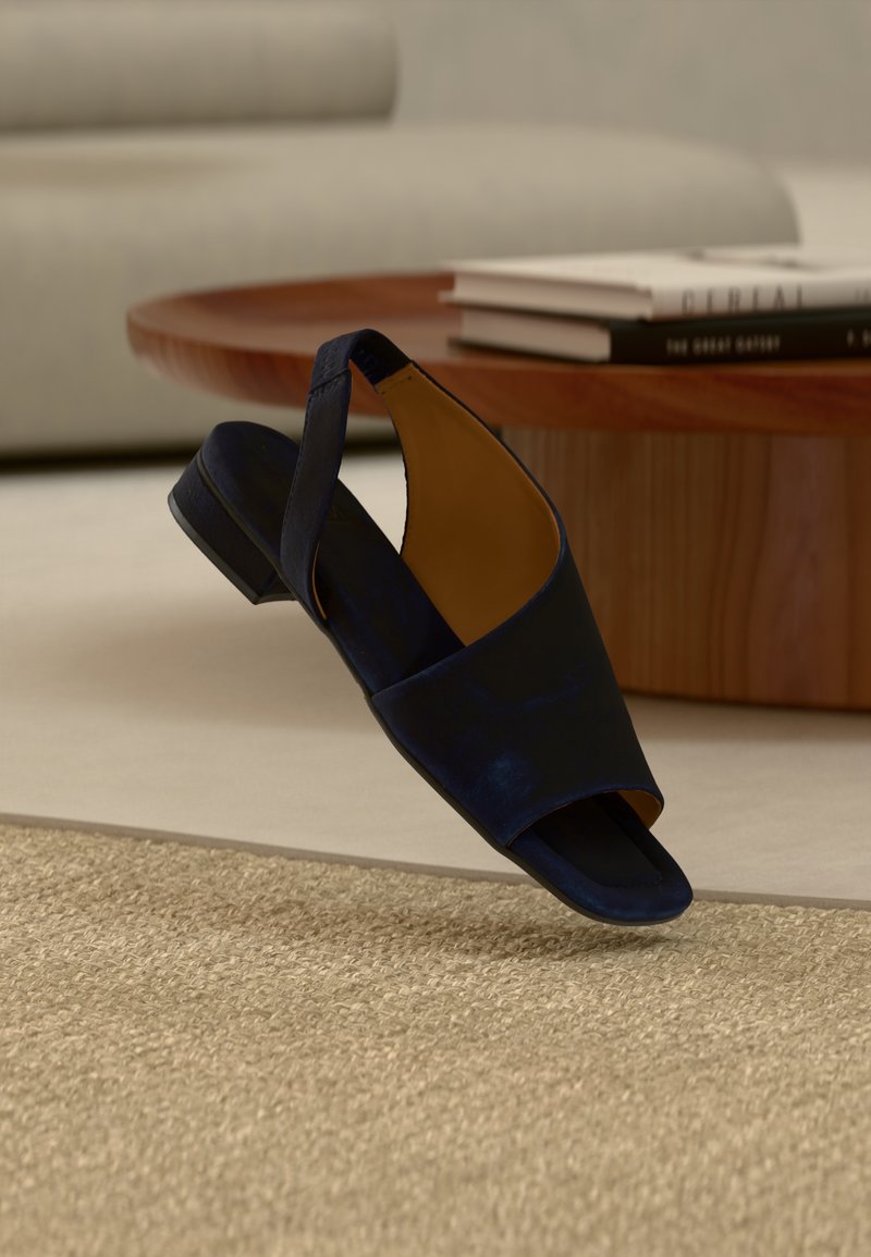 Ένα μονό πλατφόρμα ανοιχτού δαχτύλου σε ναυτικό μπλε, αιωρούμενο πάνω από ένα υφασμάτινο μπεζ χαλί, με ένα ξύλινο τραπεζάκι καφέ και στοίβες βιβλίων στο παρασκήνιο.