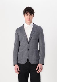 Ανδρικό γκρίζο σακάκι με υφασμάτινη υφή, δύο μπροστινές τσέπες, πέτο και κλείσιμο με ένα κουμπί, συνδυασμένο με λευκό πουκάμισο.