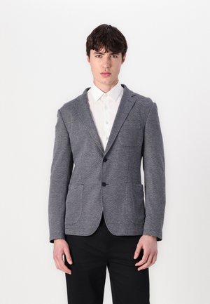 Männergrau Blazer aus strukturiertem Stoff, mit zwei vorderen Taschen, Schalkragen und einer einzigen Knopfleiste, kombiniert mit einem weißen Hemd.