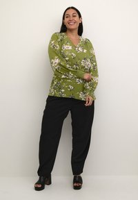 Grøn blomsterbluse med lange ærmer; har V-udskæring og smockede ærmeafslutninger. Sammen med løse sorte bukser og kraftige sorte hæle.