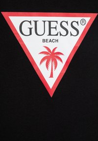 Juodas marškinėliai su dideliu baltu trikampiu raudonais kraštais. Centre esantis logotipas skelbia "GUESS BEACH" su raudonu palmės medžio grafinu.