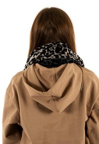 Cabello largo y castaño, lleva una sudadera color beige y una bufanda de estampado de leopardo en blanco y negro, visto de espaldas, sobre un fondo blanco.