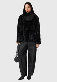 Giacca di pelliccia ecologica nera con collo alto, abbinata a pantaloni larghi in pelle nera. La modella tiene una cintura con borsa coordinata e indossa scarpe nere con punte appuntite.