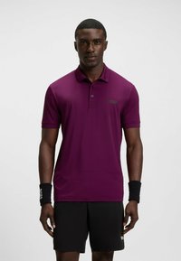 Dunkelviolettes Polo-Shirt mit kurzen Ärmeln, knöpfbarem Kragen und kontrastierendem grünen Logo. Besitzt eine glatte Textur und ein elegantes, körperbetontes Design.