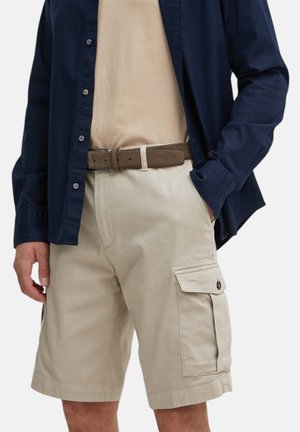 Uomo che indossa pantaloncini cargo beige con cintura marrone, t-shirt beige infilata e camicia blu navy sbottonata con una mano in tasca.