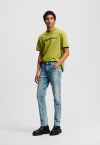 Tricou verde cu mânecă scurtă, cu imprimeu "KARL LAGERFELD JEANS", asociat cu blugi din denim albastru deschis și bocanci cu toc negru.