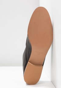 Semelle d'une chaussure en cuir noir inclinée contre un mur blanc, montrant une semelle lisse couleur fauve avec l'inscription "Made in Spain 37" en relief.