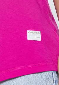 T-shirt fuchsia G-Star avec un tissu texturé, un ourlet droit et une étiquette de marque blanche avec du texte gris sur le bas.