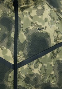 Žaliai kamufliažinė striukė, turinti tekstūruotą medžiagą, juodus akcentus, pilną užtrauktuką ir subtilų Nike logotipą krūtinės srityje.