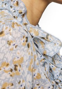 Top azul claro con estampado floral en amarillo y blanco, con escote rizado y detalles de encaje con hilos brillantes para textura y detalle.