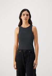 Mørkegrå ermeløs topp laget av ribbestrikket stoff, med rund halslinning og subtile tekstdetaljer foran. Kombinert med høyhalsede svarte jeans.