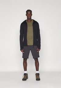 Must kapuutsiga lukuga jakk oliivrohelise T-särgi, hallide lühikeste pükste ja mustade matkasaaapadega, millel on kollased aktsendid. Valged sokid on nähtavad.