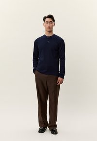 Férfi áll, sötétkék, hosszú ujjú henley ingben, barna nadrágban és fekete cipőben, kezét lazán a test mellett tartva, egyszerű háttér előtt.