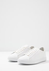 White sneakers with a smooth leather finish, round toe, flat rubber sole, and traditional lace-up design. Notable stitching accents along the sides.
