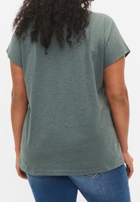 Lockig hårig person i en lös grön kortärmad skjorta och blå jeans, sedd bakifrån mot en vit bakgrund.