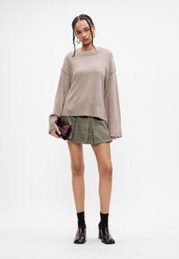 Μπεζ πλεκτό πουλόβερ με φαρδιές μανίκες, πράσινη καρο φούστα πλισέ, μαύρες κάλτσες αστράγαλου και γυαλιστερές μπορντό παπουτσιές με τακούνι. Χρυσά σκουλαρίκια.