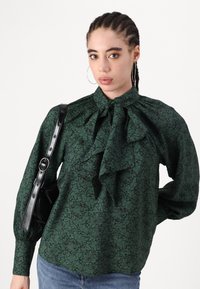 Blusa verde a fantasia con colletto alto e grande fiocco, caratterizzata da maniche lunghe a sbuffo e un tessuto testurizzato. È abbinata a una borsa nera.