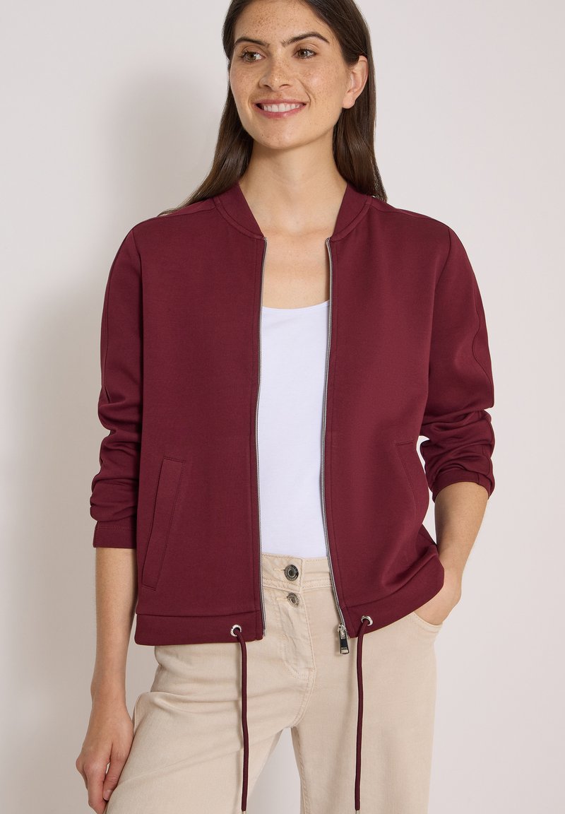 Femme souriante, portant une veste zippée bordeaux sur un haut blanc et un pantalon beige, debout avec une main dans la poche devant un fond uni.