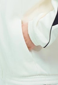 Main dans la poche d'une veste blanche avec des poignets élastiques et un détail de piping noir sur la manche.