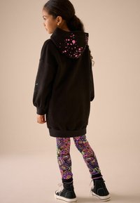 Schwarzer oversized Hoodie mit pinken Spritzern auf der Kapuze, kombiniert mit bunten Leggings mit abstrakten Mustern und schwarzen High-Top-Sneakern.