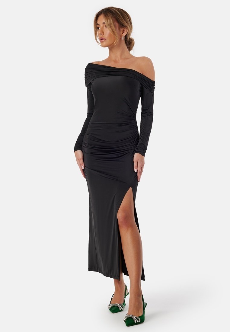 Svart off-shoulder klänning med långa ärmar, rynkdetalj, sidssprund och mjuk textur. Parat med gröna klackskor med detaljer.