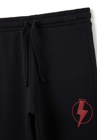 Schwarze Jogginghose mit elastischem Bund, ausgestattet mit einem roten Blitzsymbol auf dem linken Oberschenkel. Weiches Material und taillierter Schnitt.