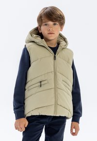 Helle khaki-Puffweste mit Kapuze, featuring Chevron-Steppung, Frontreißverschluss und Seitentaschen. Getragen über einem marineblauen Langarmshirt.